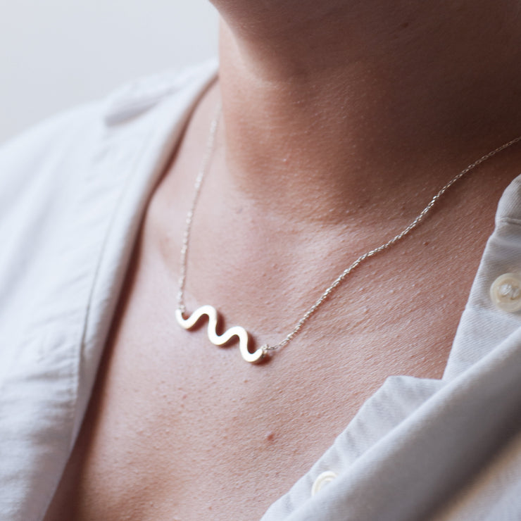 squigle line pentant set on the neck of a woman. she wear a white button down shirt that is open on the neck. 