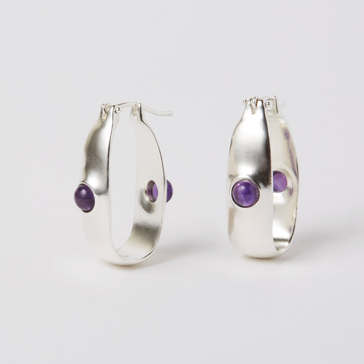 The Oval Hoop is an everyday earring, with a hint of color added by the semiprecious gem. The earrings are made of sterling silver and have a hinged ear wire made of the same material. They measure about 1.25" x 0.25". The semi precious gem is amethyst and it sits in the front and the back of the earrings.  
