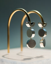 Represented are two sterling silver earrings, part of the building block collections made by Suna Bonometti. The earrings are composed of arched tops, where the ear lobe would sit. Below are a few chain links that connect with chain links two disks one below the other. The earrings are illuminated by a very bright light and are hung with brass arched stands that are set on a plexiglass platform.  The background of the image is a deep ocean blue.