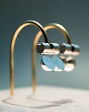 Represented are two sterling silver earrings, part of the building block collections made by Suna Bonometti. The earrings are composed of a top long cylinder, where the ear lobe would sit. Below are three disks all aligned, that are connected with chain links from the top cylinder. The earrings are illuminated by a very bright light and are hung with brass arched stands that are set on a plexiglass platform.  The background of the image is a deep ocean blue. 