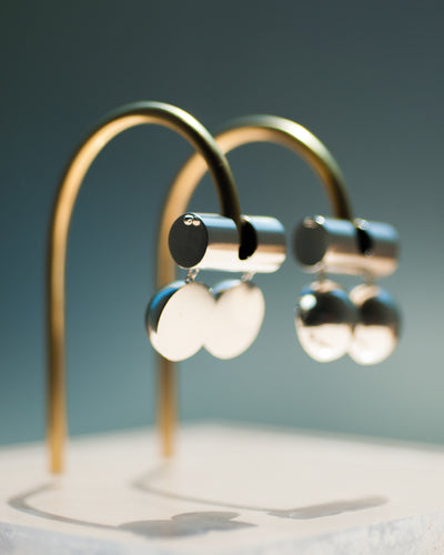 Represented are two sterling silver earrings, part of the building block collections made by Suna Bonometti. The earrings are composed of a top long cylinder, where the ear lobe would sit. Below are three disks all aligned, that are connected with chain links from the top cylinder. The earrings are illuminated by a very bright light and are hung with brass arched stands that are set on a plexiglass platform.  The background of the image is a deep ocean blue. 