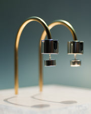 Represented are two sterling silver earrings, part of the building block collections made by Suna Bonometti. The earrings are composed of a top cylinder, where the ear lobe would sit. Below is a smaller cylinder connected to the top one by a few chain links. The earrings are illuminated by a very bright light and are hung with brass arched stands that are set on a plexiglass platform.  The background of the image is a deep ocean blue.