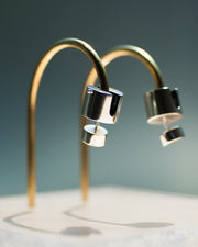 Represented are two sterling silver earrings, part of the building block collections made by Suna Bonometti. The earrings are composed of a top cylinder, where the ear lobe would sit. Below is a smaller cylinder connected to the top one by a few chain links. The earrings are illuminated by a very bright light and are hung with brass arched stands that are set on a plexiglass platform.  The background of the image is a deep ocean blue.