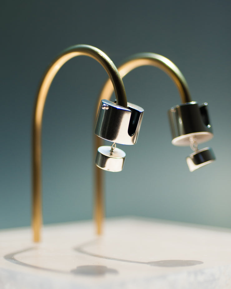 Represented are two sterling silver earrings, part of the building block collections made by Suna Bonometti. The earrings are composed of a top cylinder, where the ear lobe would sit. Below is a smaller cylinder connected to the top one by a few chain links. The earrings are illuminated by a very bright light and are hung with brass arched stands that are set on a plexiglass platform.  The background of the image is a deep ocean blue.