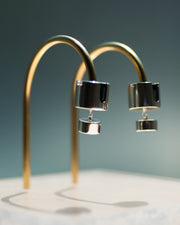 Represented are two sterling silver earrings, part of the building block collections made by Suna Bonometti. The earrings are composed of a top cylinder, where the ear lobe would sit. Below is a smaller cylinder connected to the top one by a few chain links. The earrings are illuminated by a very bright light and are hung with brass arched stands that are set on a plexiglass platform.  The background of the image is a deep ocean blue.