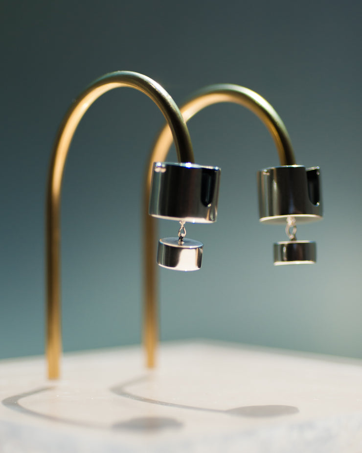 Represented are two sterling silver earrings, part of the building block collections made by Suna Bonometti. The earrings are composed of a top cylinder, where the ear lobe would sit. Below is a smaller cylinder connected to the top one by a few chain links. The earrings are illuminated by a very bright light and are hung with brass arched stands that are set on a plexiglass platform.  The background of the image is a deep ocean blue.
