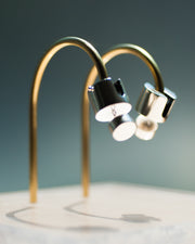 Represented are two sterling silver earrings, part of the building block collections made by Suna Bonometti. The earrings are composed of a top cylinder, where the ear lobe would sit. Below is a smaller cylinder connected to the top one by a few chain links. The earrings are illuminated by a very bright light and are hung with brass arched stands that are set on a plexiglass platform.  The background of the image is a deep ocean blue.