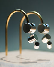 Represented are two sterling silver earrings, part of the building block collections made by Suna Bonometti. The earrings are composed of a top cylinder, where the ear lobe would sit. Below is a smaller cylinder connected to the top one by a few chain links and below that one is an even smaller one connected the same way. The earrings are illuminated by a very bright light and are hung with brass arched stands that are set on a plexiglass platform.  The background of the image is a deep ocean blue. 